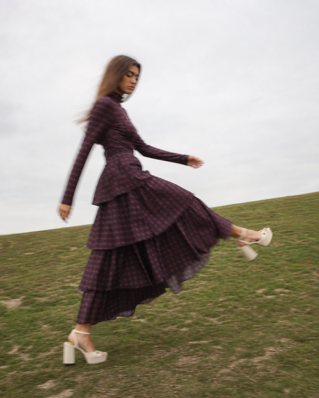 A woman wearing a dark purple and black plaid tiered maxi dress with a long-sleeve peplum bodice, captured mid-motion on a grassy hill under an overcast sky.
