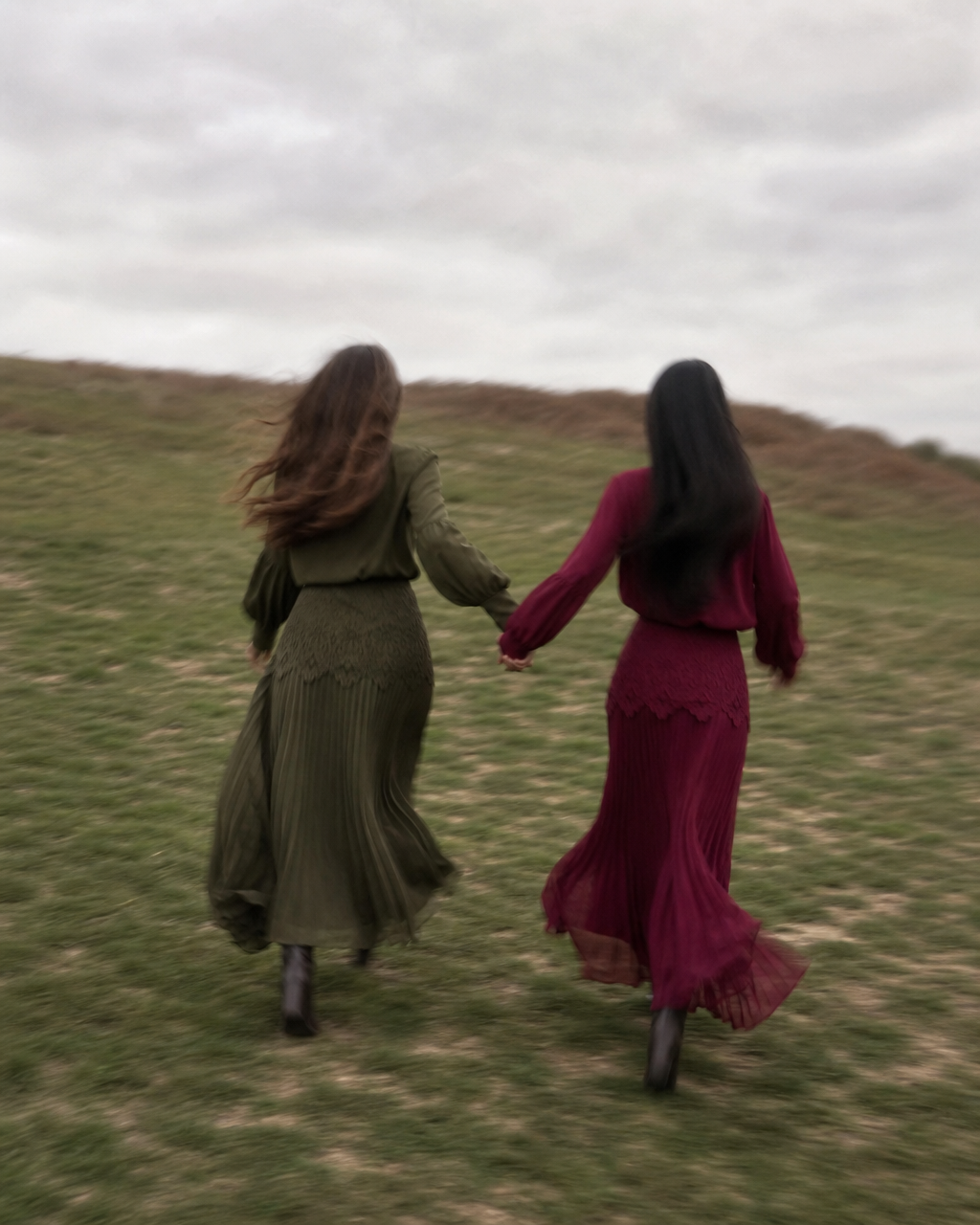 Two women holding hands and running across a grassy field under an overcast sky, viewed from behind. One wears an olive green long-sleeve maxi dress, and the other wears a deep burgundy maxi dress.